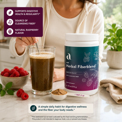 A person in a cream top stands at a kitchen counter holding a spoon in front of a glass of dark beverage, with a large tub of AIM Herbal Fiberblend supplement centered on the counter. Bowls of raspberries, a small pile of powder, and a plant are visible around the product.